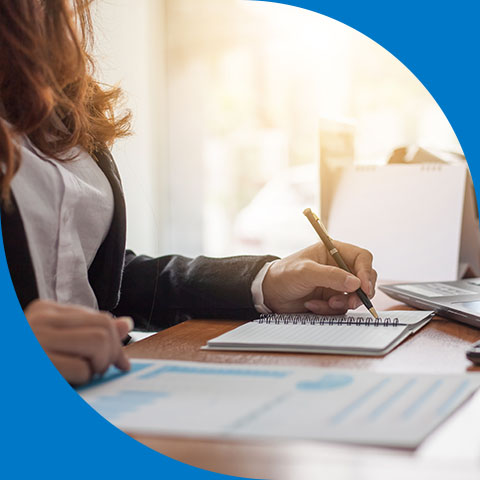 a woman sitting at a desk writing in a notepad while looking at a piece of paper next to it