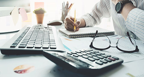 a man writing in a notepad on a desk with a calculator and pair of glasses